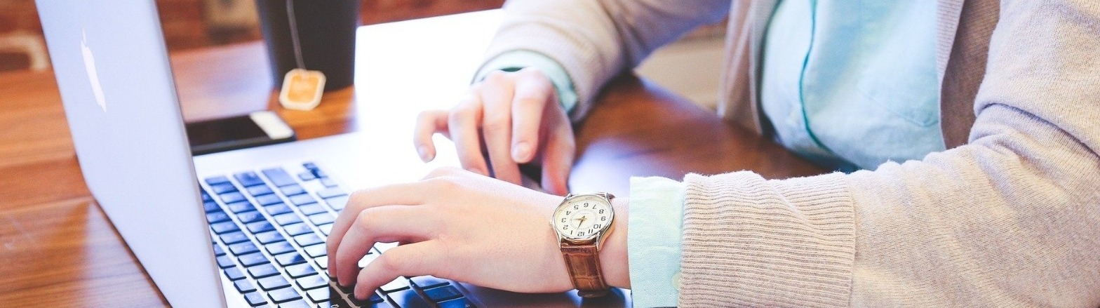 A desk with a woman's hands typing on a laptop next to her phone, cup of tea, and an open book.