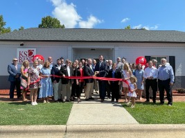 Group of people smiling in front of a building, holding a red ribbon to cut. 
