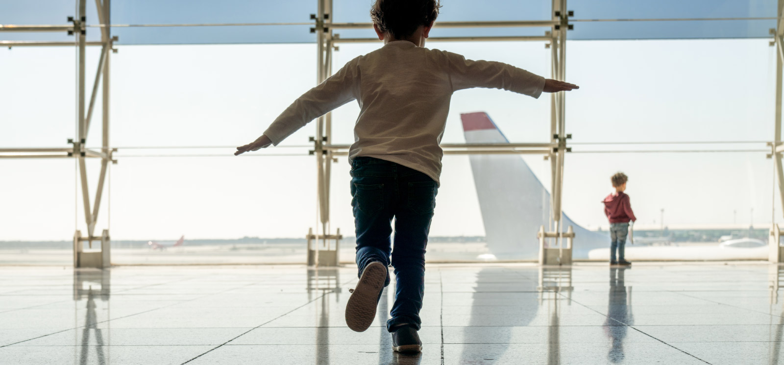 Young boy running in the airport with arms out to his side as if he is flying. While younger brother gazes out the large window admiring the planes.