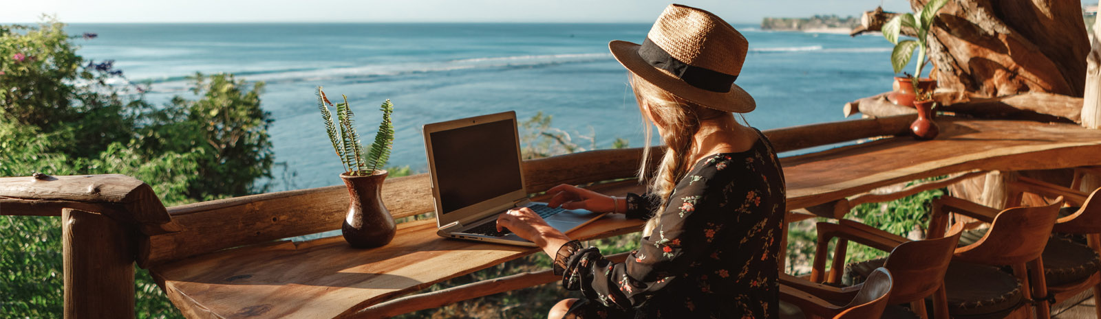 Young woman working on a laptop sits at a wooden bar overlooking the ocean.