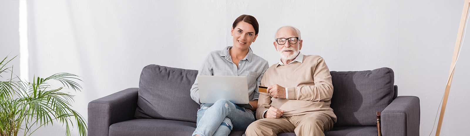 Elderly man and young woman with a laptop sit beside each other on couch.